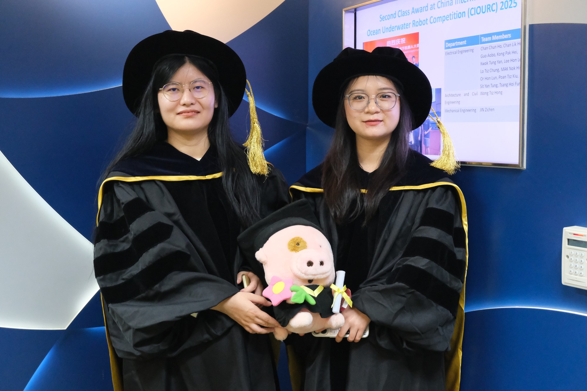 Two graduating research students in front of the Department of Electrical Engineering backdrop